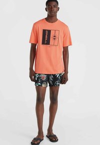 Camiseta de coral con diseño gráfico negro, combinada con pantalones cortos negros con estampado tropical colorido. Modelo lleva chancletas negras.