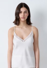 Camiseta de tirantes en suave tela color crema, con delicado encaje en el escote, tirantes ajustables estrechos y una textura suave similar a la satén.