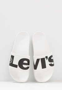 Sandalias deslizantes blancas con una banda negra gruesa que lleva "Levi's" en letras grandes. Material suave con un diseño plano y una forma ligeramente curvada.