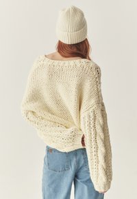 Pulover oversized de culoare crem, cu o textură groasă și modele de cabluri, asortat cu o pălărie tricotată cu model rib și blugi albaștri.