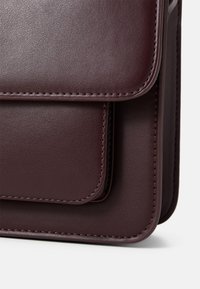 Burgunderfarget skinnhåndveske med en glatt tekstur, som har to overlappende klaffer og kontrasterende søm langs kantene.