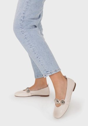 Persona que lleva vaqueros azul claro y zapatos planos color crema decorados con dos grandes cuentas plateadas en cada zapato.