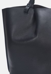 Sac fourre-tout en cuir noir avec une surface texturée, légèrement affaissé en haut, avec une bandoulière détachable et un design sans coutures.