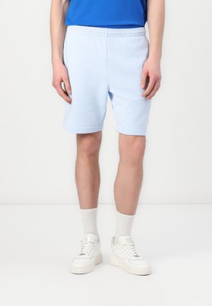 Persona che indossa pantaloncini azzurri, calze bianche corte, sneakers bianche e una maglietta blu, in piedi su uno sfondo bianco semplice.