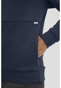 Kraljevsko modra dukserica iz mehkega materiala, z okroglim izrezom in majhno belo etiketo na spodnjem robu. Z ribastimi manšeti in robom.