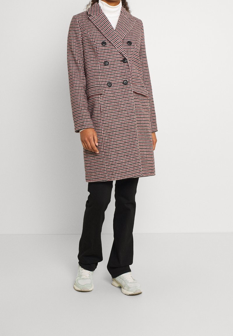 Abrigo de doble botonadura con un estampado de escocés rojo y negro, con grandes botones y un cuello, combinado con pantalones negros y zapatillas ligeras.