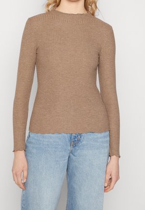 Vrouw draagt een aansluitende, geribbelde, beige trui met lange mouwen en gekartelde randen, gecombineerd met lichtblauwe, hooggetailleerde jeans, staand tegen een witte achtergrond.