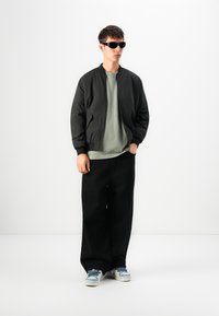 Schwarze Bomberjacke über einem hellgrünen T-Shirt, kombiniert mit weiten schwarzen Hosen und blauen Sneakers. Accessoires umfassen dunkle Sonnenbrillen.