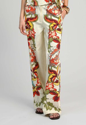 Pantalon crème à jambes larges avec grand imprimé floral orange, rouge, vert et jaune, porté avec des sandales noires et des bracelets au poignet gauche.