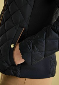 Veste noire matelassée avec un bouton-pression doré, présentant un tissu côtelé sur les côtés et un détail de poche latérale.