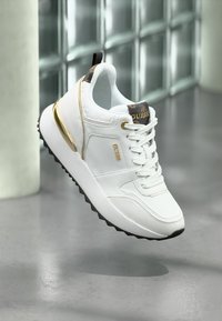 Sneaker bianca con accenti dorati, texture in pelle e tessuto, design con lacci, suola in gomma nera e logo sulla linguetta e sul lato.