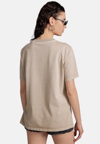 Camiseta de algodón beige de manga corta, con un corte holgado, dobladillo sin terminar y detalles de costura visibles en el cuello y las mangas.