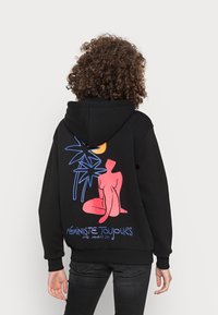 Černá mikina s kapucí s barevným grafickým designem na zádech: červená postava, modré palmové listy a žluté slunce s textem "FÉMINISTE TOUJOURS." Měkký bavlněný materiál.