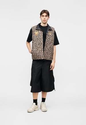 Joven con un chaleco de estampado de leopardo sobre una camiseta negra, pantalones cortos anchos negros, calcetines negros y zapatillas beige, de pie frente a un fondo liso.