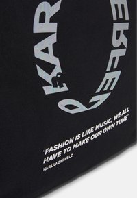 Svart handväska med vit text; innehåller en grafisk design med "KARL" och en figur, tillsammans med ett citat av Karl Lagerfeld.