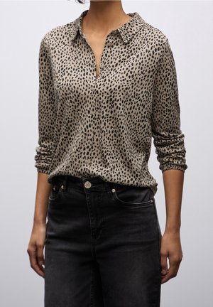 Femme portant une chemise à manches longues beige à imprimé léopard, boutonnée et rentrée dans un jean en denim noir taille haute, manches retroussées.