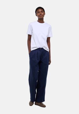 Femme portant un t-shirt blanc uni à manches courtes, un pantalon ample bleu marine avec plis et des chaussures plates à motifs, debout les mains dans les poches.