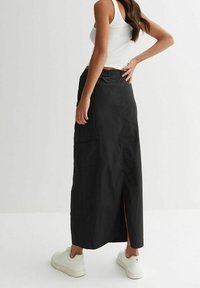 Vrouw draagt een zwarte lange rok met een zijzak en een split aan de achterkant, een wit tanktop en witte sneakers, terwijl ze met een hand op haar heup staat.