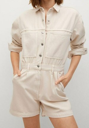 Nainen, jolla on päällään kermaisen värinen napillinen haalari, jossa on kääritty hihat, etutaskut ja shortsit, kädet sivutaskuissa.