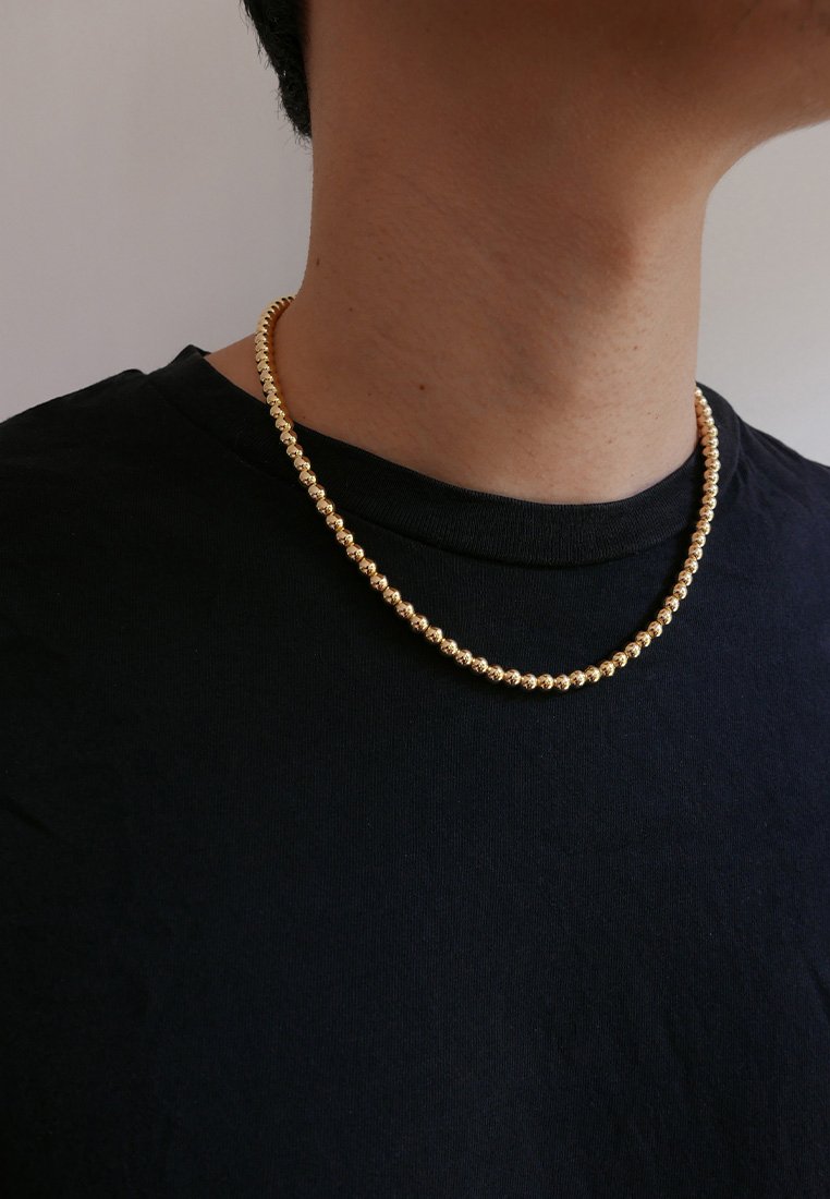 Zlata perla ogrlica, prikazana na vratu osebe, s uniformnimi okroglimi biseri z sijočo površino, nošena čez črno majico.