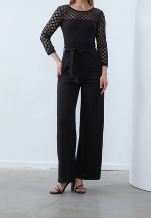 Vrouw die een zwarte jumpsuit draagt met kanten driekwartmouwen, een ceintuur in de taille, broek met wijde pijpen en zwarte sandalen met hak.