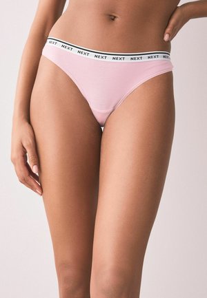 Kvinde iført lyserødt undertøj med hvid elastisk linning, hvor teksten "NEXT" gentages, hånd hvilende ved hoften.