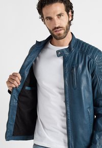Blauwe leren jas met ritssluiting, geribbelde schouderdetails en een borstzak, open gedragen over een wit t-shirt. Glad textuur.