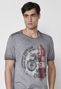 Camiseta de algodón gris con un gráfico de motocicleta vintage en negro y rojo. Detalles desgastados y un escote clássico tipo crew. Mangas cortas.
