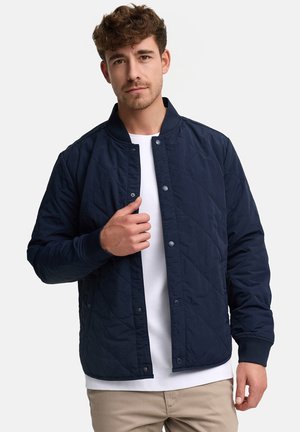 Mies, jolla on kiharat ruskeat hiukset, pukeutunut laivastonsiniseen tikattuun takkiin valkoisen paidan ja beigejen housujen päällä, napittaa takin kiinni.