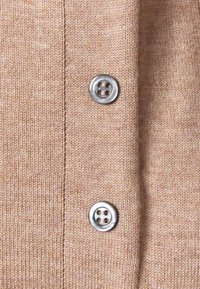 Gebreide cardigan in lichtbruin met twee metalen knopen en fijne ribstructuur; heeft een centrale sluiting die langs de voorkant loopt.