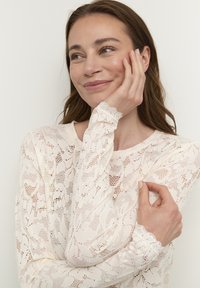 Top in pizzo a maniche lunghe in avorio con motivi floreali, polsini ondulati e scollatura rotonda, realizzato in un tessuto morbido e testurizzato.