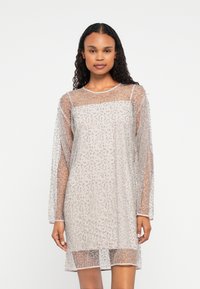 Egy térd alatt érő, áttetsző bézs ruhát visel�ő nő, hosszú ujjakkal, amelyet apró ezüst gyöngyök díszítenek, egy sima fehér háttér előtt áll.