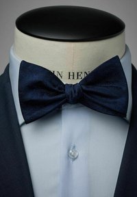 Mörkblå fluga med en strukturerad mönstring, bärs över en ljusblå skjorta under en mörk jacka, med en tydlig knappstängning.