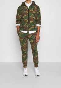 Conjunto de sudadera con capucha y pantalones con patrón de camuflaje en verde y marrón, con cremallera frontal y capucha. Combinado con zapatillas blancas y una camisa con cuello debajo.