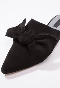 Mule en daim noir avec un design à bout pointu, ornée d'un grand nœud à l'avant et d'une doublure intérieure lisse.