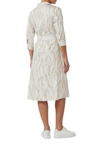 Weißes Kleid mit beigem Blattmuster, Kragen, Dreiviertelärmeln und Taillenkordel, kombiniert mit weißen Turnschuhen. Glatte Stoffstruktur.