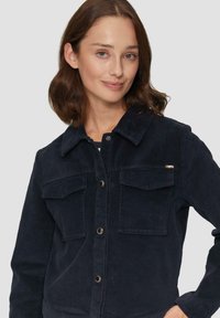 Granatowa kurtka z korduroy z zapięciem na guziki z przodu, dwoma kieszeniami na piersi i kołnierzykiem. Teksturowany materiał z widocznymi żeberkami.