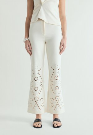 Mujer con pantalones blancos acampanados con patrones simétricos cortados con láser por encima de los tobillos, combinados con top blanco, pulsera plateada y sandalias negras.