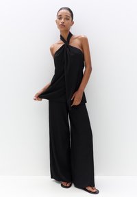 Crni halter top od teksturirane tkanine, uparen s crnim hlačama širokih nogavica. Outfit sadrži uvijen dekolte i opuštenu siluetu.