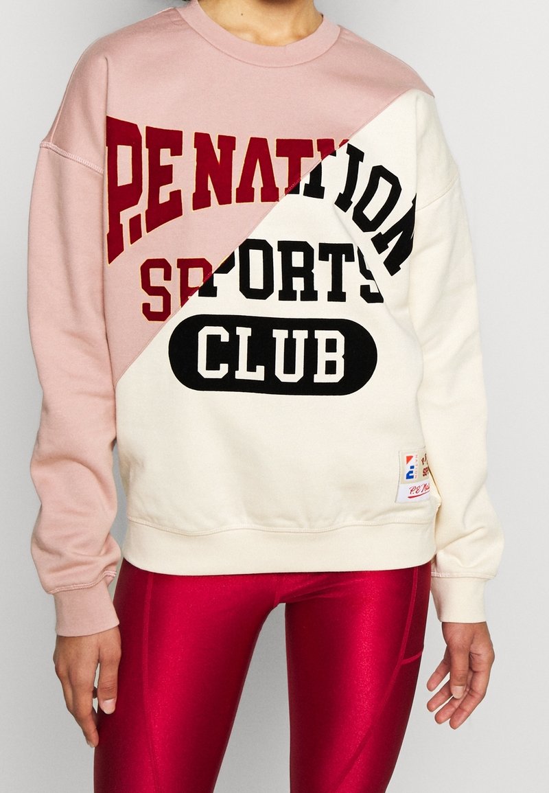 Personne portant un sweat bicolore rose et blanc avec le texte « Penation Sports Club » et des leggings rouges brillants, visible du cou jusqu’aux hanches.