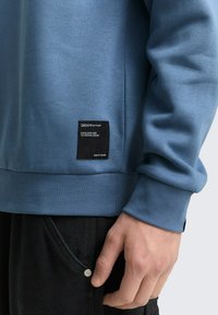 Niebieska bluza z ściągaczami na mankietach i dolnym hemie, z czarną tkaniną naszywaną z boku. W zestawieniu z czarnymi dżinsami, które mają guzik.