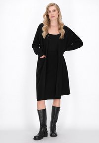 Svart lång cardigan med ribbat mönster, oversized passform och framfickor, ihop med en figurnära svart klänning och knähöga svarta stövlar.