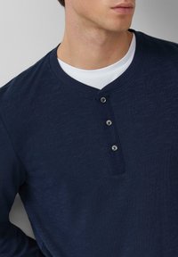 Man in een marineblauwe henley-shirt over een witte crewneck t-shirt, met zicht op het bovenlichaam en de nek, kijkt naar rechts.