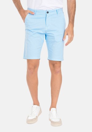 Uomo che indossa pantaloncini celesti fino al ginocchio e scarpe da ginnastica bianche, in piedi con una mano in tasca davanti a uno sfondo semplice.