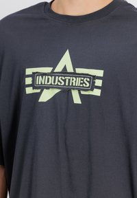 Sötétszürke, rövid ujjú póló, amelyen egy nagy világoszöld logó látható, az "INDUSTRIES" felirattal fekete téglalap alakú háttéren.