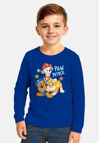 Camicetta azzurra a maniche lunghe con cani dei cartoni animati di "Paw Patrol." La grafica include testo e stelle colorate, abbinata a pantaloni grigi con impronte di zampe.