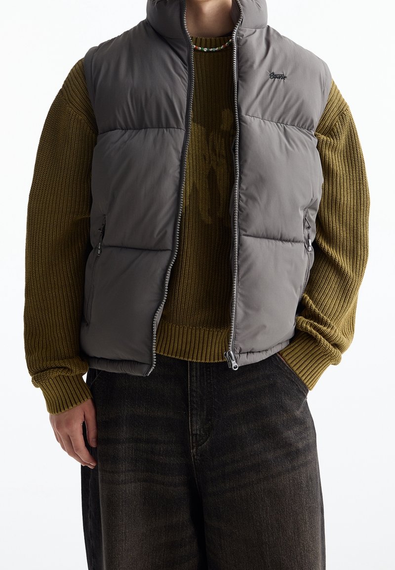 Personne portant un pull en maille vert olive, un gilet matelassé gris avec fermeture éclair, un jean foncé et un collier de perles, debout avec une main dans la poche.