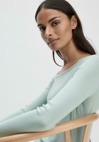 Maglietta verde chiaro a maniche lunghe con scollatura tonda, realizzata in tessuto morbido. Presenta una texture liscia e un design aderente.
