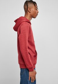 Felpa rossa in cotone con tasca frontale, cordini bianchi e polsini a costine. Indossata sopra jeans di denim blu, mostrata di lato.