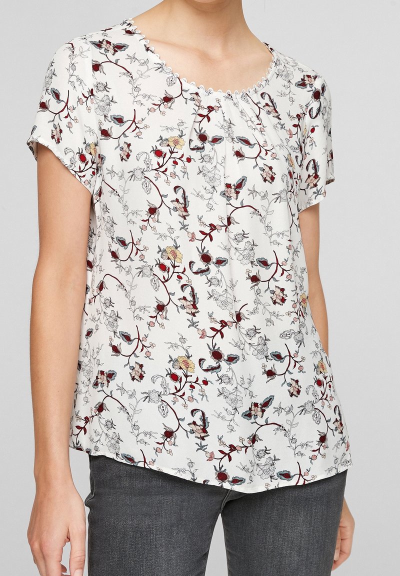 Femme portant un chemisier blanc à manches courtes avec des motifs floraux rouges, jaunes et bleus et un pantalon gris foncé.